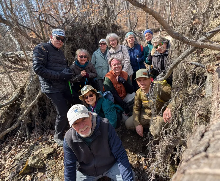 A group of around 11 people outdoors in a forest. They are all standing or kneeling between the bases of two large fallen trees. They are all dressed for cooler weather. Some are wearing hats and glasses. They are all smiling.