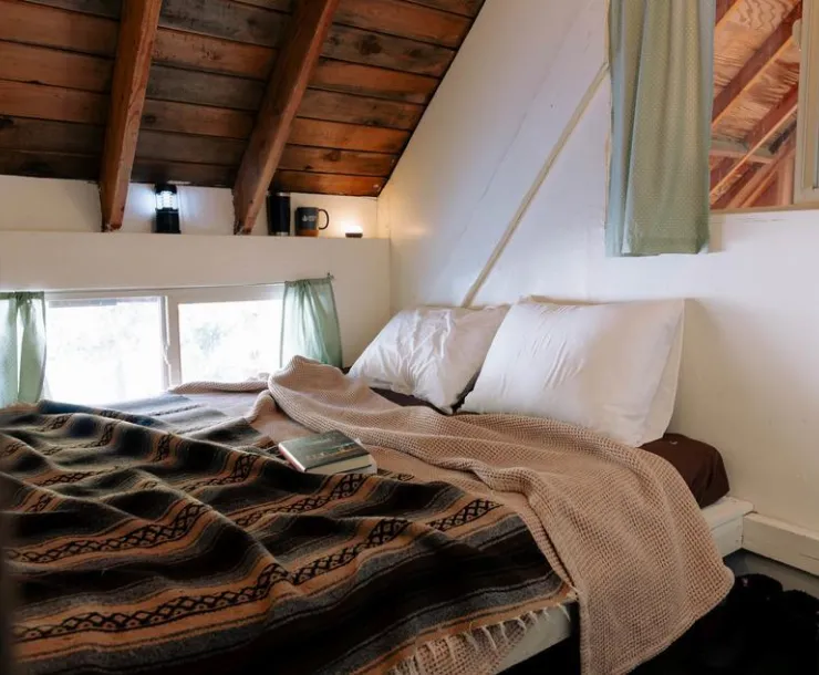 A room with a slanted wooden ceiling, white walls, and a cozy looking bed with blankets