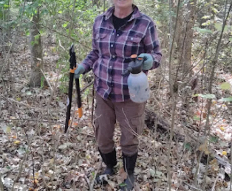 A person in the woods with a spray bottle in one hand and a tool for cutting weeds in the other. She is wearing a light blue baseball cap, long pants, knee-length boots, a checkered shirt, and blue work gloves. She is smiling. There are some green leaves left on the trees and lots of brown leaves on the ground.