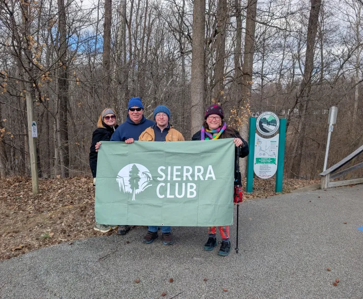 Four people at a trail head holding a green Sierra Club banner. They are dressed for the cold weather in coats and hats. The trees behind them are bare and there are brown leaves on the ground.