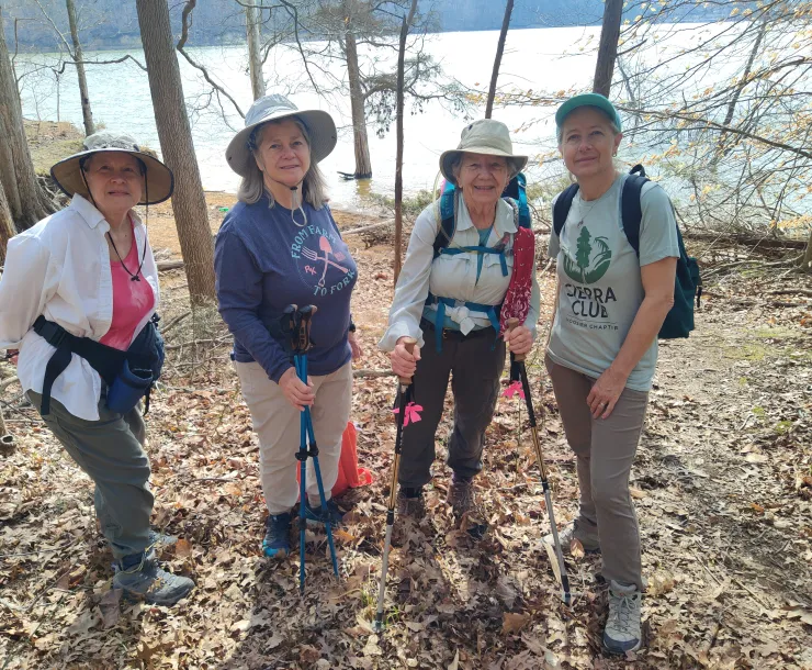 Four people on a hike with a body of water behind them and sparse trees with brown leaves on the ground. They are wearing sun hats and some of them have hiking poles. One of them is wearing a Sierra Club tee.
