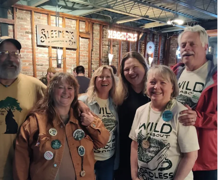 A group of six people indoors at a bar with exposed brick walls. A few of the people are wearing tees that say Wild About Roadless Forests. They are all smiling.