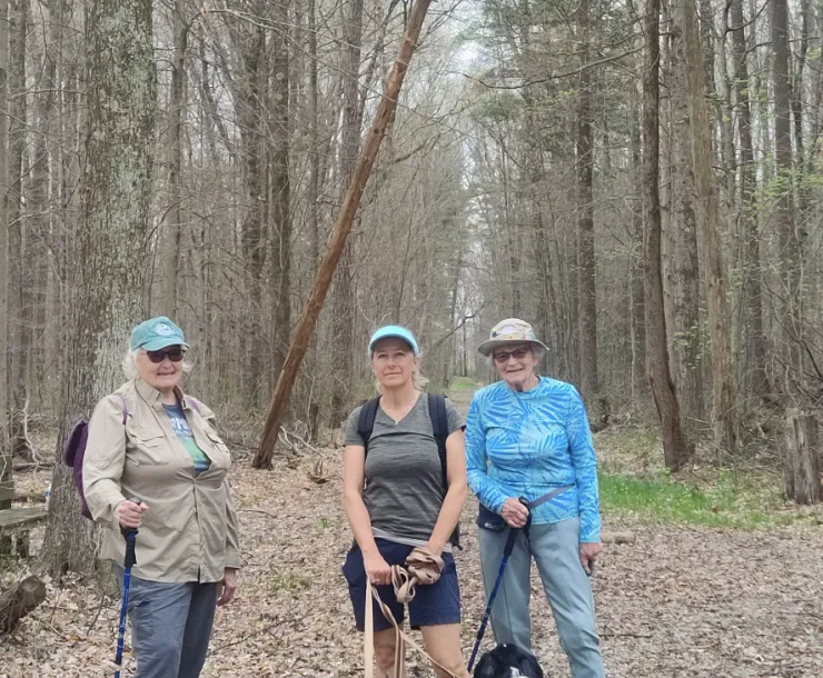 Three people on a leafy trail with tall trees behind them, with two black and white dogs. They are all smiling. 