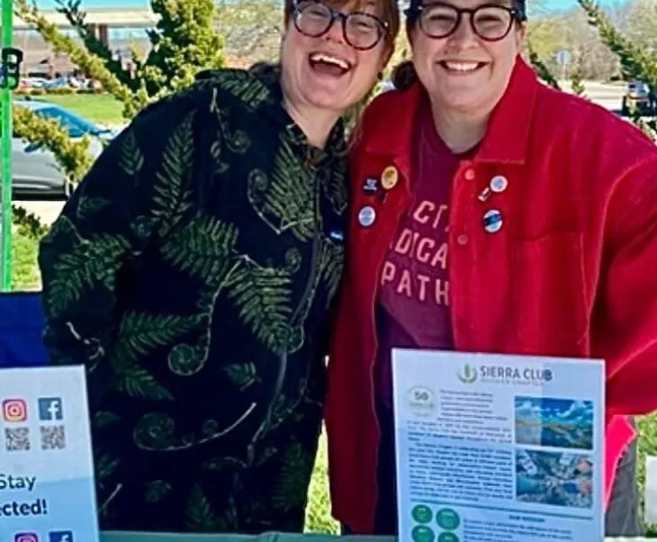 Two people tabling at an event outdoors. The sky is blue behind them. There is a green tablecloth on the table and information about Sierra Club. They are both wearing glasses and are smiling joyously.