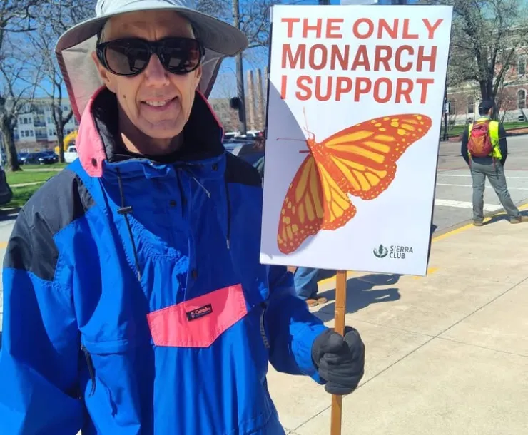 A person in a blue coat and a sun hat and dark shades, holding a sign saying The Only Monarch I Support with an illustration of an orange and red butterfly, and a Sierra Club logo in the corner. They are standing near a road by a park, and the sky is blue.