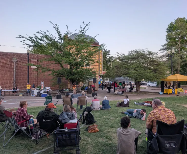 Several people outdoors at an event. There are colorful tents set up with tables including one where people can register to vote. The grass is green and the sky is blue. 