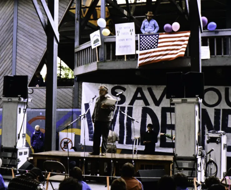 a man, Harvey Manning, stands on a stage in front of a crowd with a large black and white hand painted banner behind him that reads "save our wilderness"