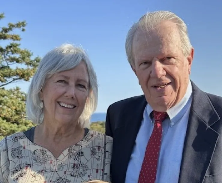 Head and shoulders photo of two people outdoors with a hazy blue sky behind them and some green trees in the background to the left. The person on the right has short gray hair and is wearing a blue suit and red tie. The person on the left has mid length gray hair and a pale top with a dark red pattern. They are both smiling.