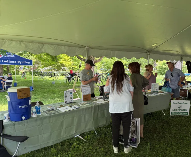 Five people under a large tent at a tabling event. Two of them are behind the table talking to folks. On the table are flyers and other information about Sierra Club, and a blue water cooler with a sign on it saying Visit With Sierra Club!