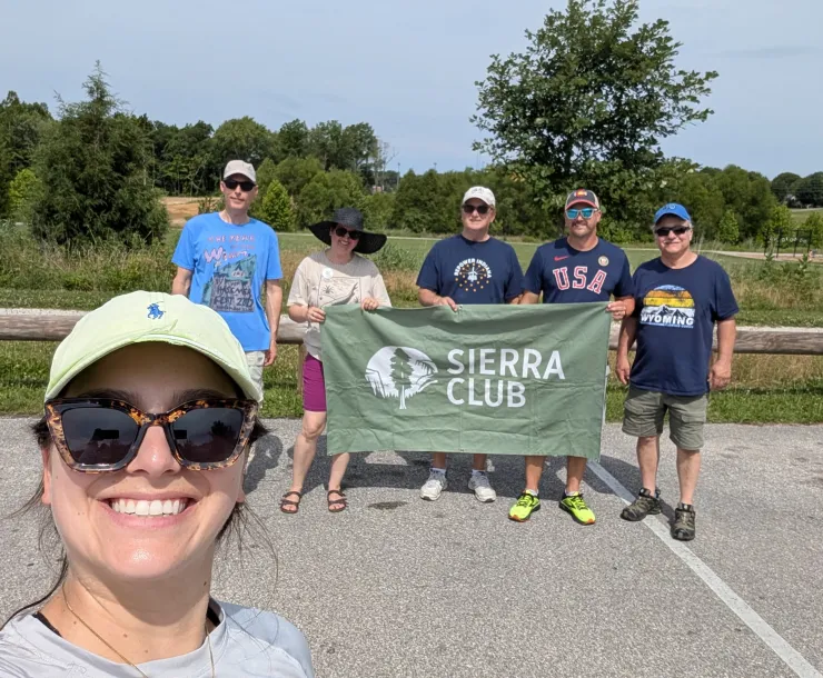 Five people in a parking lot with a park behind them with trails and trees and a hazy blue sky. The three folks in the middle are holding a green Sierra Club banner. In the foreground is the person taking the photo, selfie-style. She has a baseball cap and sunglasses and is smiling.
