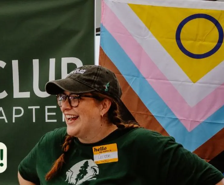 A head and shoulders photo of a person wearing a green Sierra Club t-shirt, a black cap, and with a Sierra Club Hoosier Chapter and a Pride flag behind them. They are outdoors tabling and are smiling and looking off to the side. The text says Happy Pride Month and there is a Sierra Club Hoosier Chapter 