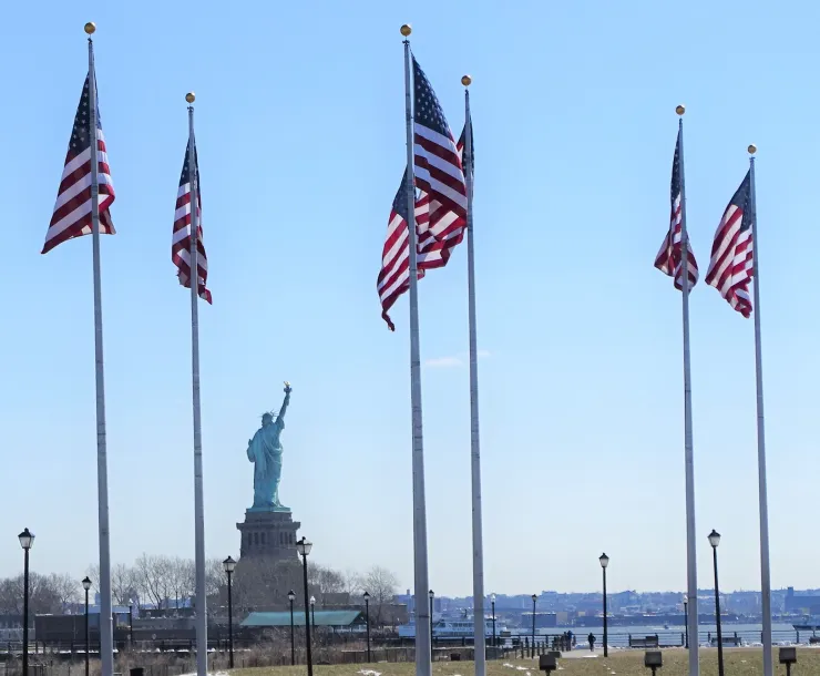 American flags wave in the foreground with a view of the Statue of Liberty looking over the Hudson River.