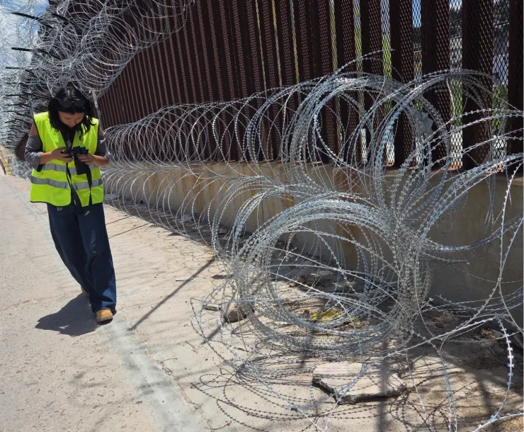 A person in a yellow vest stands by the border wall next to a pile of concertina wire