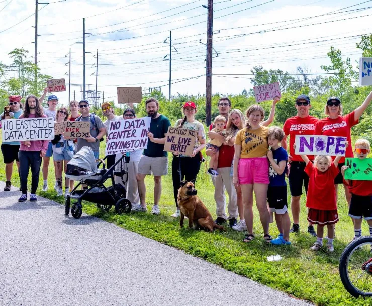 A group of around 20 people on a city trail. There is a paved trail path with grass to the side. They are holding signs to protest against a data center. A couple of them have bicycles. They are a mix of ages including some small children.