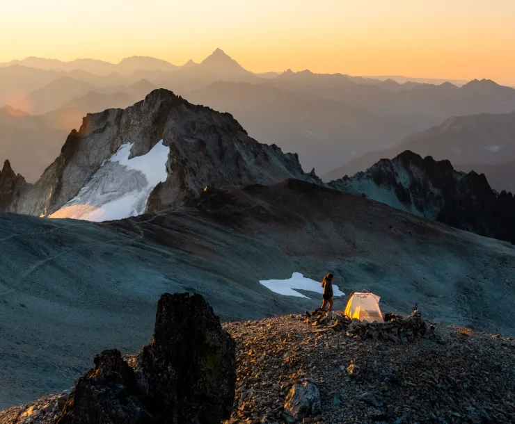 Layers of jagged mountain peaks, with a few small snowfields, under a soft yellow-orange sunset. There is a person with a tent perched on a small flat area in the foreground. 
