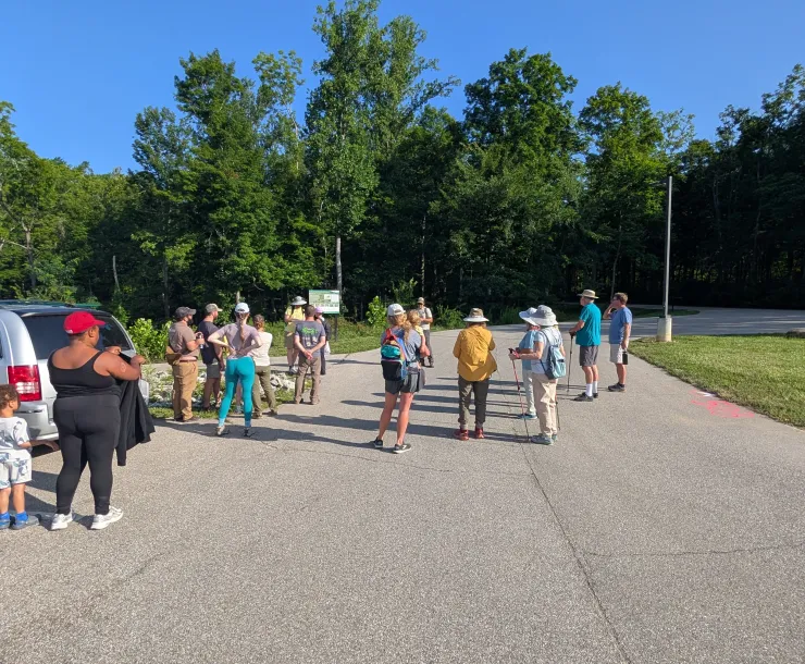 Around sixteen people in a parking lot by a trailhead. Many are wearing caps, and some have hiking polls. They are all facing away from the camera, listening to a person talking.