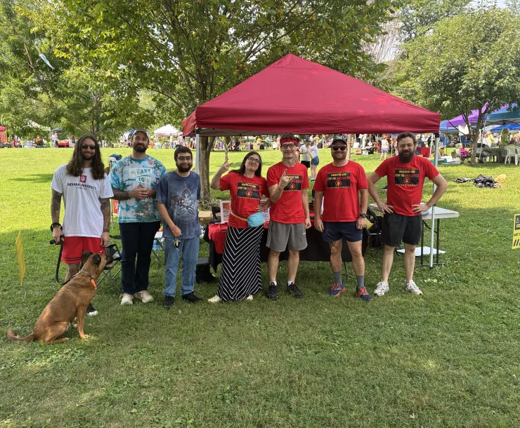 Seven people and a dog standing in front of a tabling tent in a park with trees and other tabling tents in the background, with folks milling around. Four of the people in the photo are wearing red t-shirts which say Irvington says Stop the Data Center