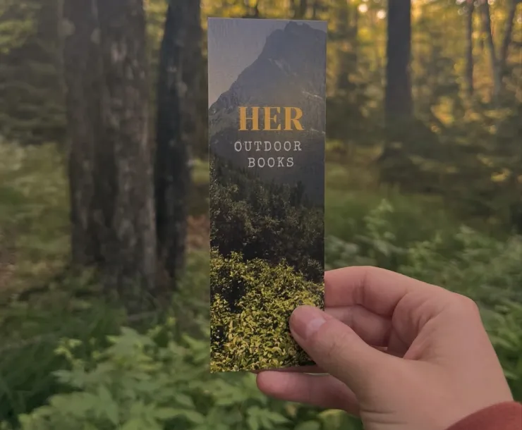 Photo of a hand holding a Her Outdoor Books bookmark in a sunlit forest