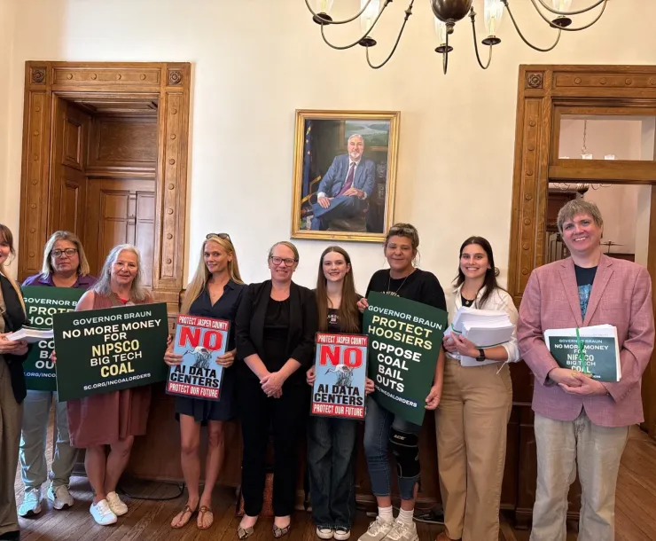 Nine people standing indoors, holding signs saying things like No AI data centers. Protect Hoosiers - oppose coal bail outs. Some of them are holding stacks of paper petitions.
