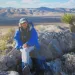 An older woman sitting on a rock in the desert.