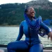 A still from the film The Black Mermaid, featuring a Black woman in a purple wetsuit with long purple braids out on the water, living life to the fullest. 