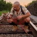 Alexi Grousis squats down in the middle of some train tracks. He has a metal hook tool and is holding up what looks like an old turtle shell.