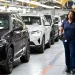 A female worker in all blue walks by a line of new cars in a factory.