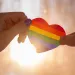 Hand hold a heart painted like a LGBT flag, silhouetted against sun