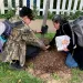 Citlally Fabela and Dulce Garduno squat down to examine the mulch around a recently planted tree. A white picket fence is in the background.