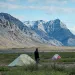 A man stands near four tents in an Arctic Refuge valley meadow with snowcapped mountains in the background.