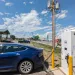A royal-blue Tesla is charging at an Otterspace station with a sign that reads "Electric Vehicle Charging Station" next to a store and a road.