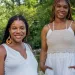 Diamon Clark and Kristen Walker stand by a tree-lined creek wearing long white dresses.