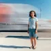 Alicia Rivera is wearing a light blue shirt, jean shorts, and flip flops and standing in front of traffic by the port.