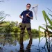 Dick Jordan smiles at the camera and holds a net while wearing tall wading boots and standing in a grass-lined body of water.