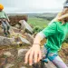 Two kids wearing harnesses and helmets hike over rocks at the top of a hill.
