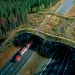 Aerial photo shows two two-lane highways and a curved grassy overpass surrounded by dark-green trees on both sides. A large truck with a red top is coming through a tunnel.