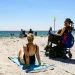 A person sits in a wheelchair on the beach, next to another person sitting on a towel on the sand.