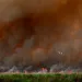 A fire burns across a green field and brown smoke fills the sky.