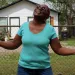 Sandra Edwards wears a turquoise shirt and sunglasses and holds her hands up to the sky and smiles. Behind her is a chain-link fence and a small white house.