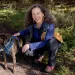  Julia Olson wears hiking clothes and smiles while kneeling down with her brown dog on a trail.