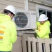 Two workers in hard hats and neon yellow hoodies install a heat pump on top of a home's beige siding 