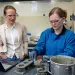 Leah Ellis wears a light blazer and white button-down shirt, watching Summer Camerlo-Bass in a blue work shirt scooping gray cement from a metal mixing bowl.