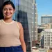 Laura Zapata stands against a glass wall inside a skyscraper with a city in the background