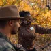 A Black woman in camo shoots a bow and arrow among fall foliage