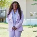 Catherine Coleman Flowers wears a lavender pantsuit and stands in front of a house, smiling at the camera.