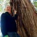 Rick Lanman stands with his left hand on a redwood tree trunk as he looks down at the camera.