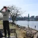 A man stands on a bench near a river, pointing binoculars toward the sky. 