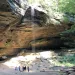 Sunlight pours through the trees onto a wide-angle view of Ash Cave with a trickle of water falling 