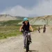 Miriam Peterson wears a turquoise helmet and rides on an unpaved road toward the camera with two other riders and the gray, rocky Oregon Buttes behind her.