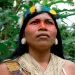 Close-up of Nemonte Nenquimo looking into the distance, wearing a feather headdress and string top. A red stripe is painted across her eyes and nose.